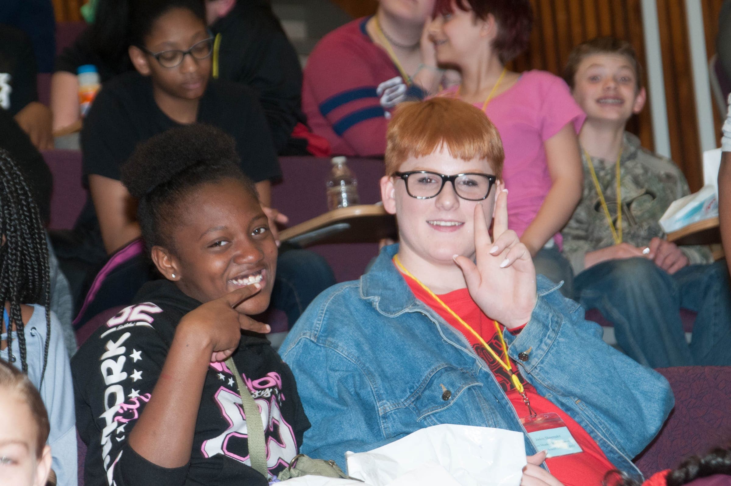 Two students grinning at the camera in a lecture hall during the AAYLC conference, one flashing a peace sign, with classmates wearing lanyards behind them